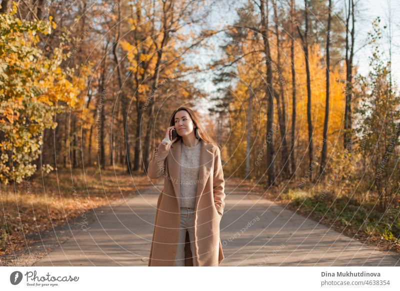 Junge brünette Frau mit langen Haaren in beige Mantel zu Fuß im Herbst Park auf einer Straße. Entspannende Momente in der Natur, Millennial Mädchen glücklich zu sein und lächelnd. Frau spricht am Telefon zu Fuß im Herbst Park.