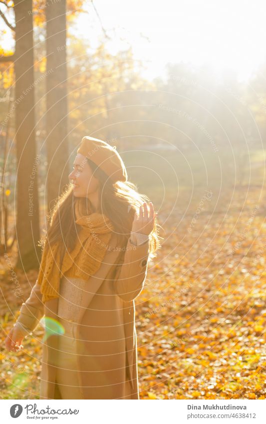 Junge Frau in gelber Baskenmütze und Schal und in beige Mantel zu Fuß im Herbst Park bei Sonnenuntergang mit warmen Sonnenschein. Entspannende Momente in der Natur, Millennial Mädchen glücklich zu sein und lächelnd.