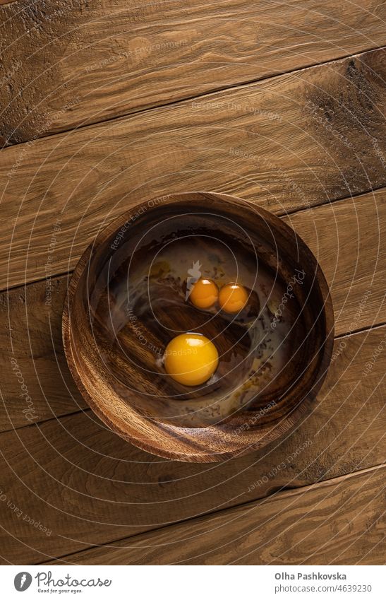 Runde Holzschale mit zerbrochenen rohen Hühnereiern. Double Dotter Ei und gewöhnliche Ei aussehen wie lustiges Gesicht mit Augen und Nase. Natürliche braune Holztisch Hintergrund. Unmittelbar über Ansicht. Platz zum Kopieren.