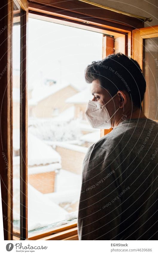 Junger Mann mit Maske in Quarantäne, der durch das Fenster seines Hauses den Schneefall betrachtet allein Angst begrenzt Einsperrung Kontemplation Korona