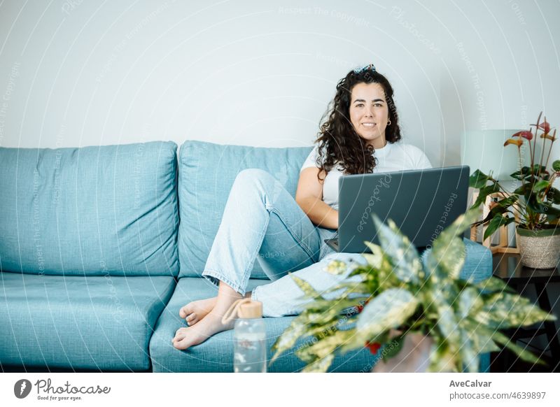 Junge Freiberufler Latina Frau arbeitet am Computer, während auf dem bequemen Sofa zu Hause auf moderne trendige Kleidung sitzen. Konzept der Remote-Arbeit von zu Hause aus, junge Menschen online arbeitet