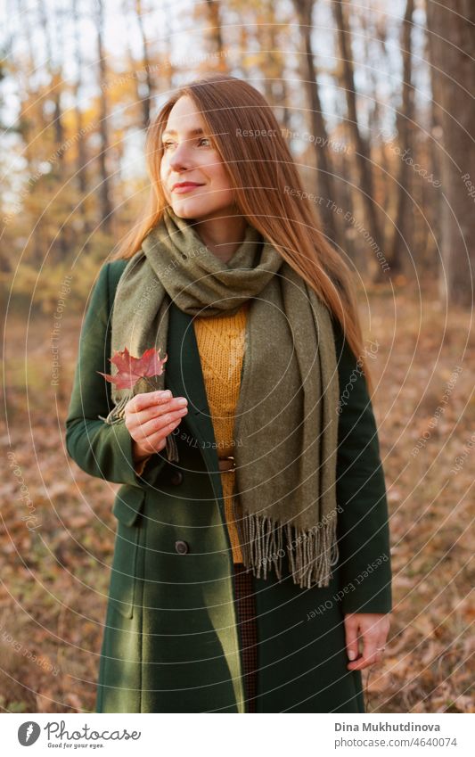 Schöne Frau mit langen Haaren in grünem Mantel steht im Herbst Park hält einen Herbst Ahornblatt. Stil, Mode und Kleidung. Tief in Gedanken im Herbst Wald in der Natur. Authentischer Lebensstil.