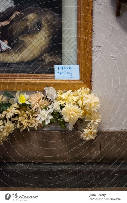 Seltsame Kombination aus künstlichen Blumen, einer Ecke eines Gemäldes und einem handgeschriebenen Schild mit der Aufschrift "think spring". künstliche Blumen