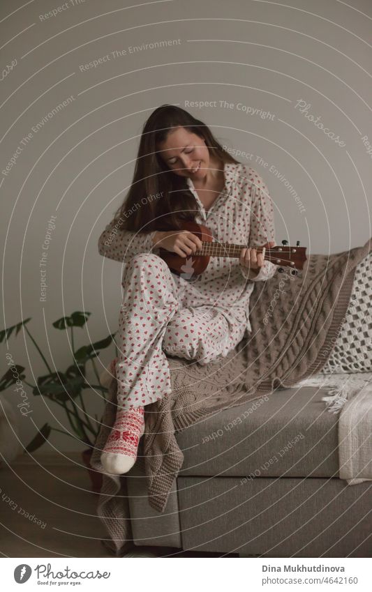 Junge Frau zu Hause in Pyjamas spielen Gitarre sitzen auf einer Couch oder Sofa. Millennial weiblichen Musiker in gemütlichen Wohnung spielen Ukulele in Urlaub Weihnachtszeit Pyjamas gekleidet. Talentierte junge Person übt Ukulele spielen.