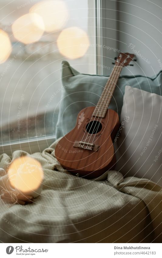Ukulele zu Hause auf der Fensterbank mit kuscheligen Decken und Kissen an einem kalten Regentag. Gemütliche Stimmung, die Zeit zu Hause verbringen. Melodie und Musik Hobby - kleine Gitarre zum Musizieren.