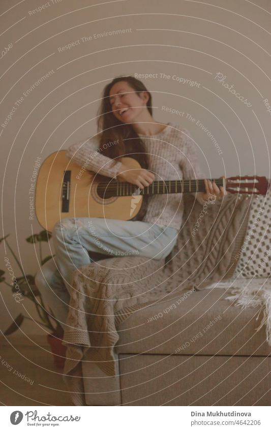Glückliche junge Frau spielt Gitarre und singt, sitzt auf einer Couch in gemütlichen Zimmer zu Hause in der modernen Wohnung. Musik und Hobby zu Hause oder auf einer Party mit Freunden.
