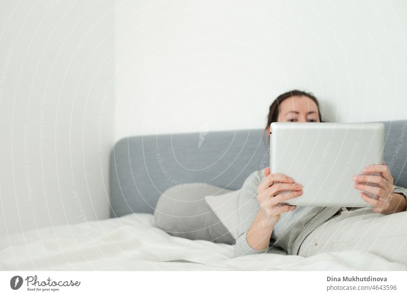 Junge Frau arbeitet im Home-Office mit Tablet auf dem Bett liegend. Schaut Filme oder arbeitet online. Informelles Büro zu Hause. Graue Farbpalette Bild der Person mit Technologie mobiles Gerät.