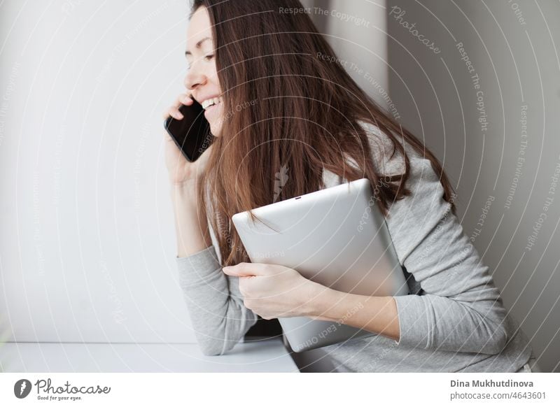 Junge Millennial-Frau mit braunen Haaren spricht am Handy und hält Tablet-Gerät. Graue Farbpalette. Frau arbeitet im Büro oder zu Hause hält mobile Geräte. Communicationa bei der Arbeit und Online-Geschäft Konzept.