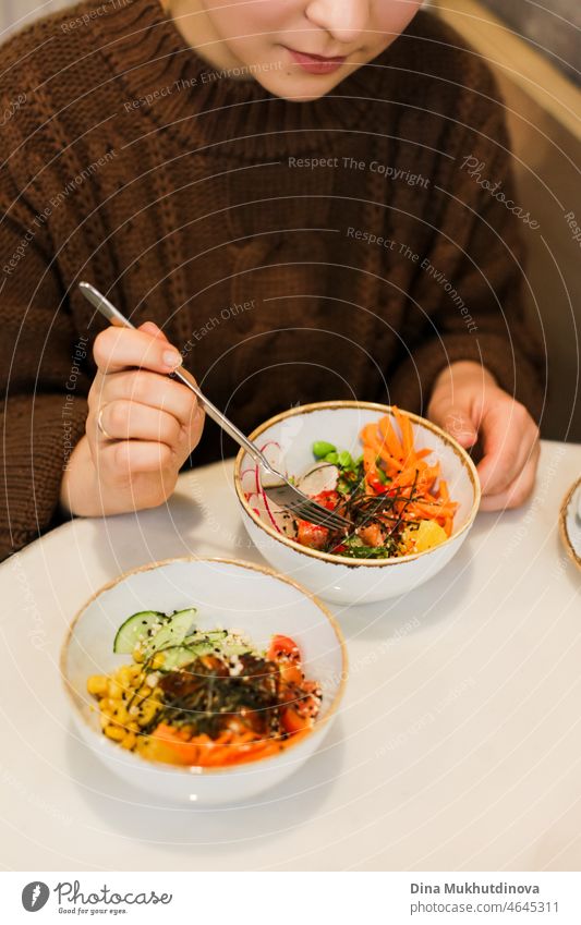 Frau im braunen Pullover isst eine Poke Bowl oder Salat im Restaurant. Buntes gesundes Essen - Gemüse und Fisch und Meeresfrüchte. hawaiianisch Hawaii Overhead