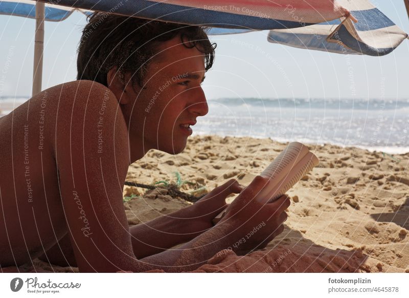 Strandleben: Jugendlicher schaut beim Lesen auf das Meer lesen Buch Lektüre Junge Teenager Sonnenschirm Lesestoff Roman Urlaub entspannt Sand Urlaubsstimmung