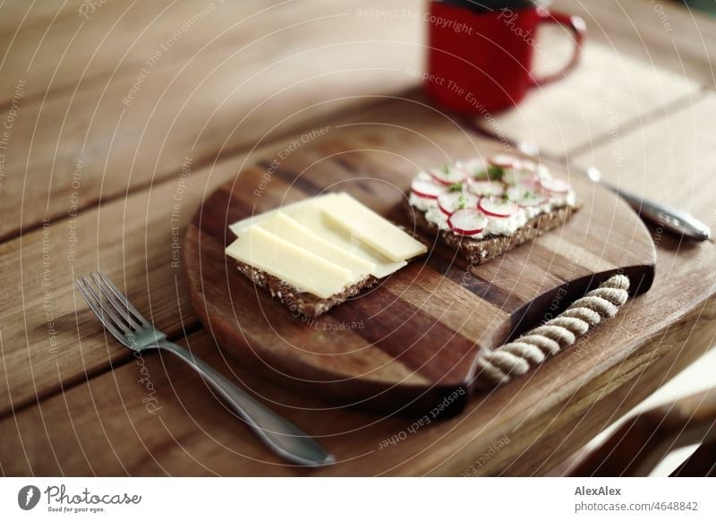 Gesundes Frühstück - eine Scheibe Schwarzbrot belegt mit Gruyère und eine mit körnigem Frischkäse, Radieschen und Kresse - auf einem Frühstücksbrettchen aus Holz mit Besteck auf dem Holztisch mit Kaffee in einer roten Tasse
