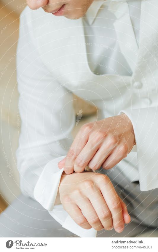Bräutigam im weißen Smoking sitzt und sein Hemd feststeckt striegeln brünett Mann verheiratet Hochzeitsmorgen formal Kleidung Hand Schaltfläche Feier Anzug Mode