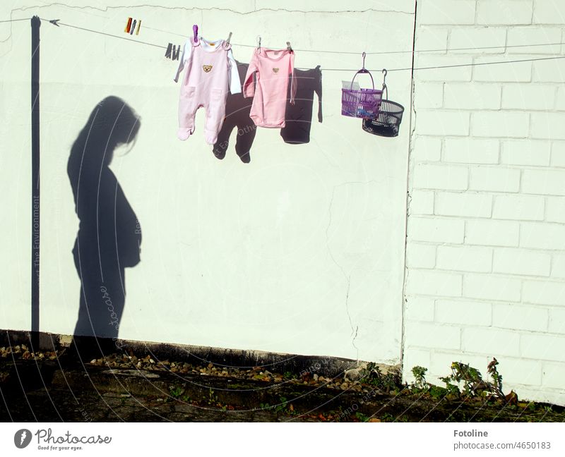 In freudiger Erwartung - Auf einer Wäscheleine hängt frisch gewaschene Babykleidung und ein Körbchen mit Klammern. Der Schatten der werdenden Mutter zeigt... lange dauert es nicht mehr, bis das Baby kommt.