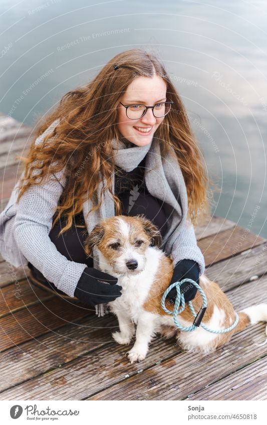 Eine junge Frau mit Brille und langen Haaren sitzt mit einem kleinen Hund auf einem Holzsteg. Terrier Haustier Tier niedlich Außenaufnahme Lifestyle