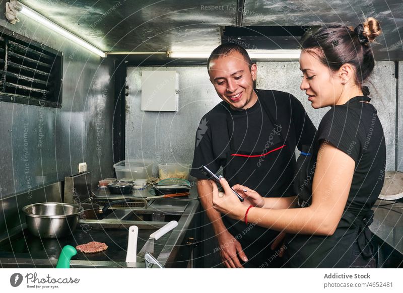 Positive Foodtruck-Besitzer teilen Smartphone Mann Frau braten Kotelett Imbisswagen Straßenessen benutzend Kleinunternehmen Burger Pastetchen Koch Funktelefon