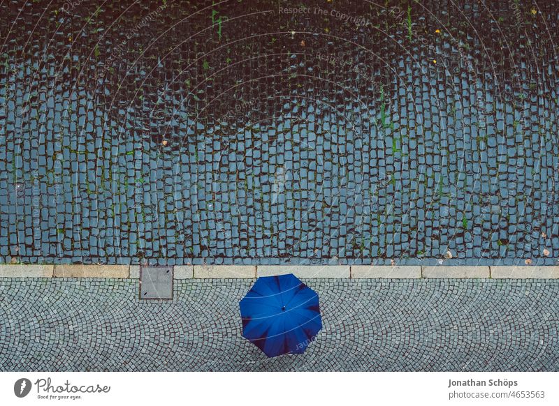 Vogelperspektive auf Person mit Regenschirm auf Straße bei Regen nass Pflastersteine gepflastert Fußweg schlechtes Wetter Regenschutz grafisch minimalistisch