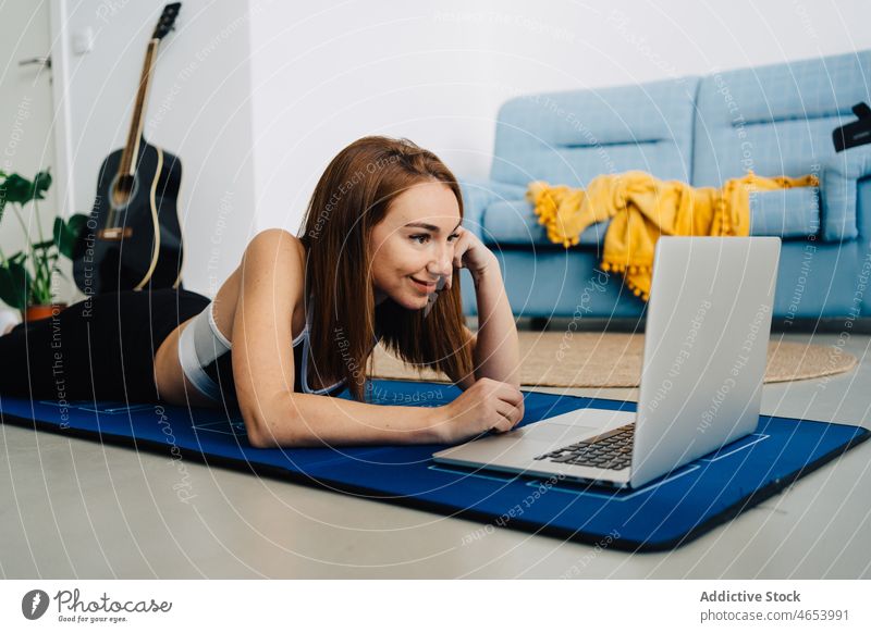Lächelnde Frau auf Matte liegend mit Laptop üben benutzend Yoga online Fitness zuschauen Video Tutorial Netbook jung Unterlage Internet Training Sportbekleidung