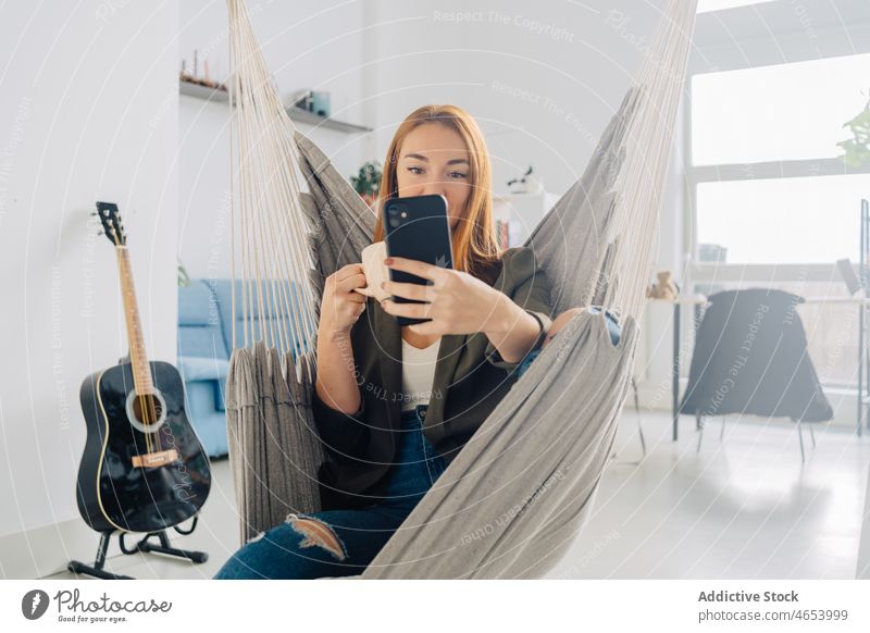 Frau nimmt Selfie in Hängematte mit Kaffee Smartphone benutzend Selbstportrait Kälte sich[Akk] entspannen Freizeit Heißgetränk zu Hause trinken ruhen rote Haare