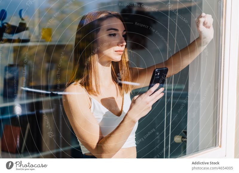 Junge Frau mit Smartphone am Fenster benutzend Mobile Internet nachdenklich Anschluss online Browsen jung Telefon soziale Netzwerke Surfen Kommunizieren