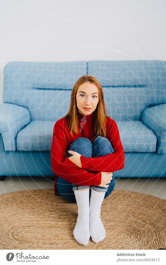 Frau umarmt Knie, während sie in der Nähe des Sofas sitzt Knie umarmend Liege Teppich schüchtern ruhen einsam Rotschopf unsicher sitzen Komfort rote Haare