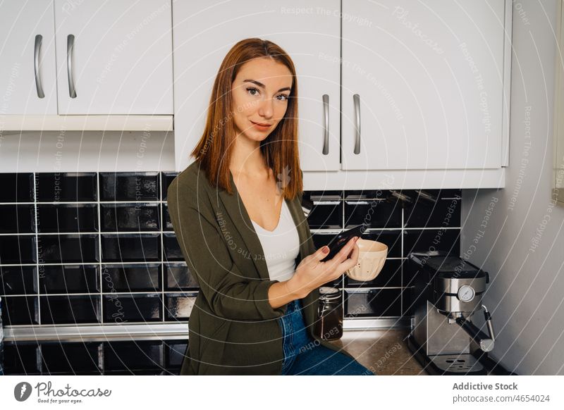 Frau mit Tasse Heißgetränk und Handy in der Küche Smartphone Funktelefon benutzend Abfertigungsschalter trinken Kaffee Texten Konzentration sitzen Jeansstoff
