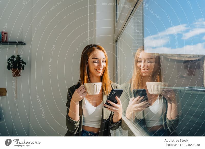 Fröhliche Dame mit einer Tasse Kaffee, die in der Nähe des Fensters auf ihrem Smartphone surft Frau Funktelefon heiter benutzend trinken Heißgetränk Lächeln