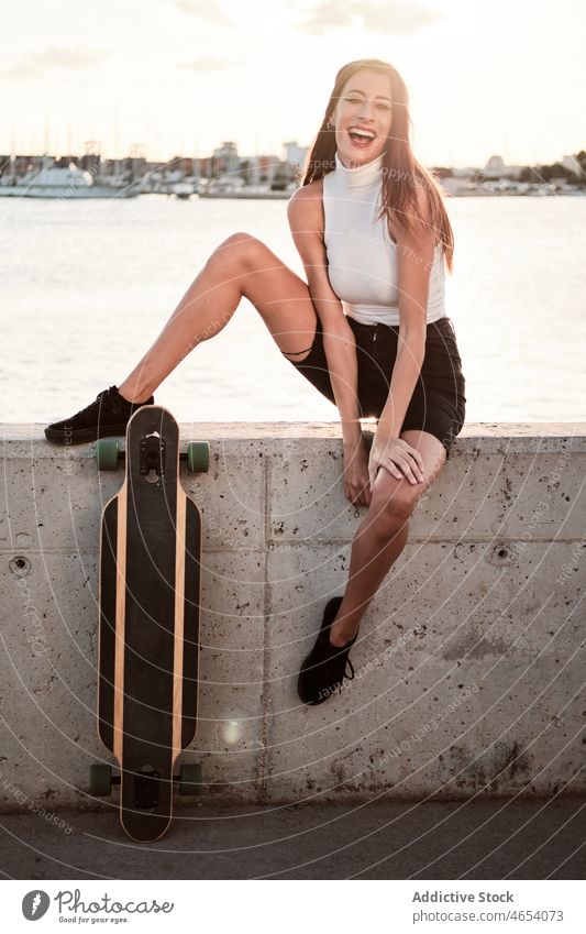 Lächelnde Frau mit Longboard auf einer Böschung MEER Stauanlage Sommer Stil sich[Akk] entspannen Wochenende Freizeit Vorschein heiter schlank Meeresufer jung