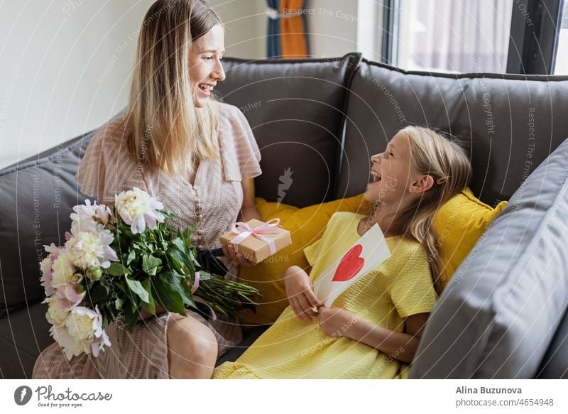 Kind Tochter gratuliert Mutter und gibt Geschenkkarte, Geschenk und Blumenstrauß zu Hause. Mama und Mädchen lächelnd und umarmt auf der Couch. Glückliche Mütter Tag Feier