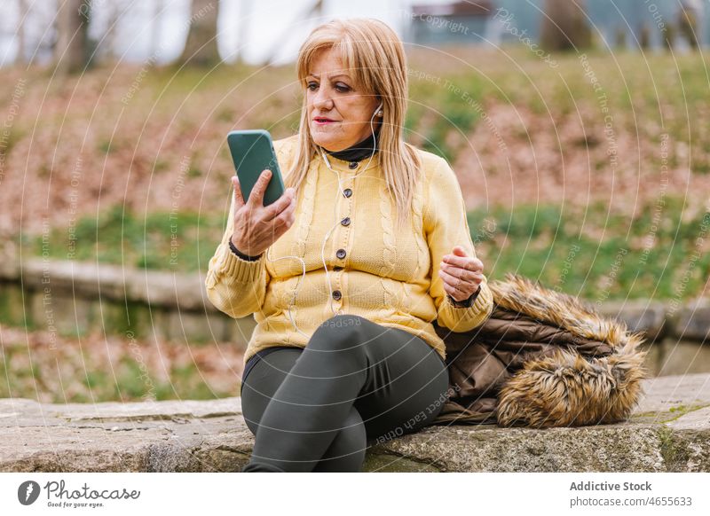 Ältere Frau, die im Park auf ihrem Smartphone surft Kopfhörer Musik zuhören Browsen Textnachricht online Internet Talkrunde Lebensmitte Anschluss Vorschein