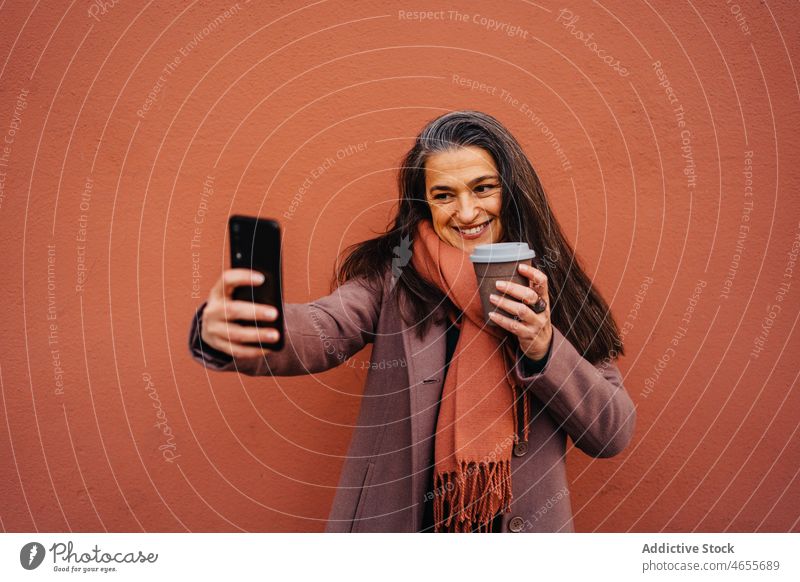 Positive reife Frau trinkt Kaffee und macht ein Selfie mit ihrem Smartphone Funktelefon Selbstportrait Imbissbude benutzend zum Mitnehmen Lächeln Telefon Mobile