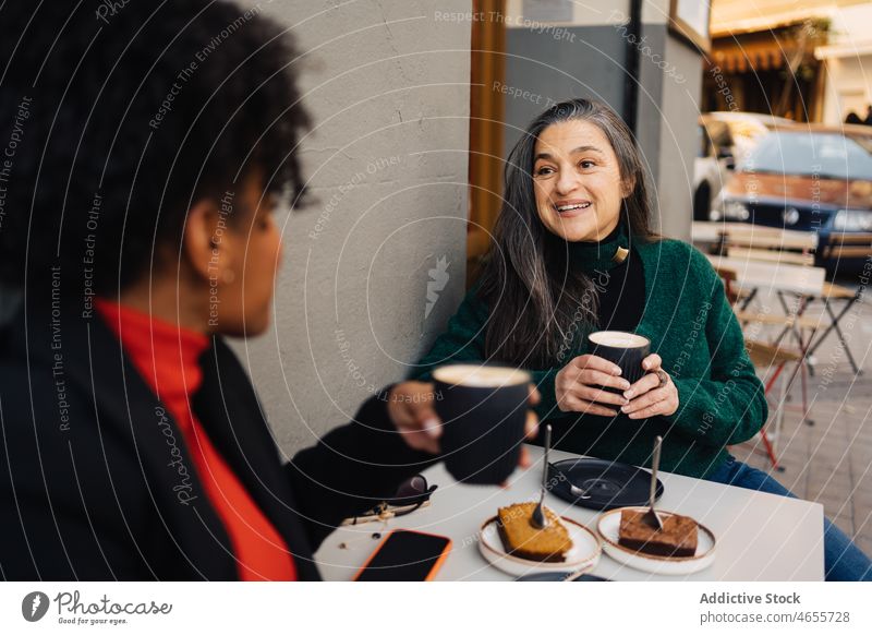 Positive ethnische Frau trinkt Kaffee und unterhält sich mit einem schwarzen Freund Frauen Latte Sitzung Dessert Kuchen Kaffeehaus Talkrunde positiv reden