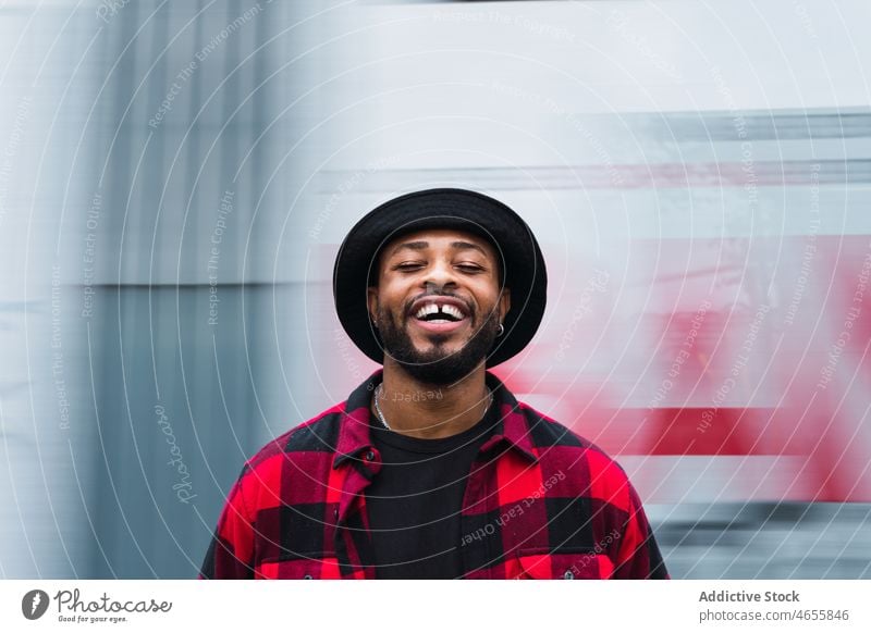 Glücklicher ethnischer Mann auf verschwommener Straße heiter Hut Lächeln Vorschein Lachen froh Persönlichkeit Stil Porträt männlich positiv Individualität