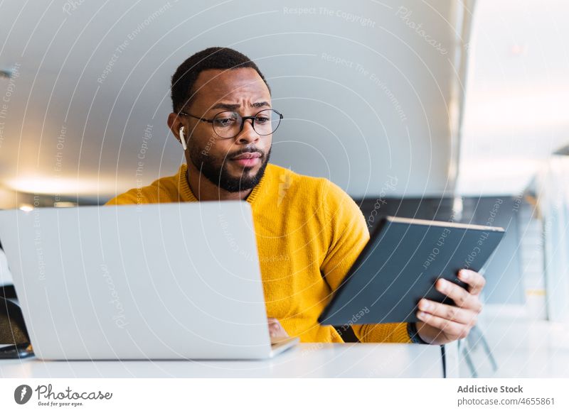 Mann mit Ohrstöpseln, der ein Tablet und einen Laptop am Tisch benutzt Tablette Kopfhörer benutzend nachdenklich zuhören Musik Arbeitsplatz meloman ernst