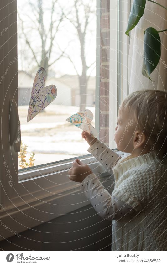 Kleinkind hängt handgefertigte Papierherzen in das Fenster ihres Hauses; selbstgemachte Valentinstagsdekoration Herz Dekorationen diy Valentinsgruß Handwerk