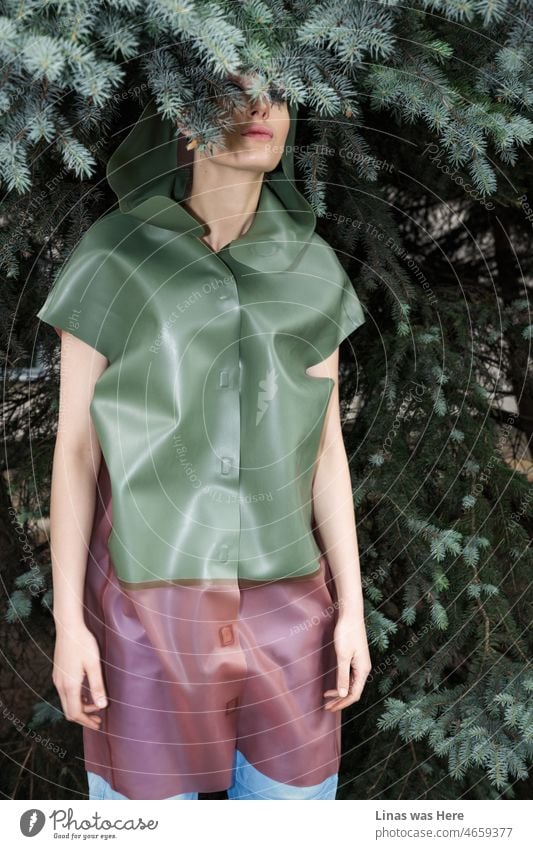 Ein in einen Latex-Regenmantel gekleidetes Mädchen versteckt sich in diesen Büschen. Tannenbäume mit grünen Ästen passen perfekt zu ihr auf diesem Bild. Ein Modemodell zeigt die besten Eigenschaften von Latex auf eine andere Art und Weise.