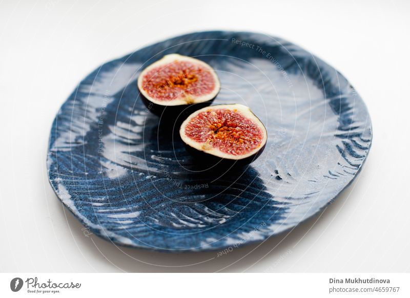 Eine reife Feige in zwei Hälften geschnitten auf einem blauen handwerklichen Teller auf weißem Tisch. Köstliche reife Feigenfrucht als Dessert für eine gesunde Ernährung.