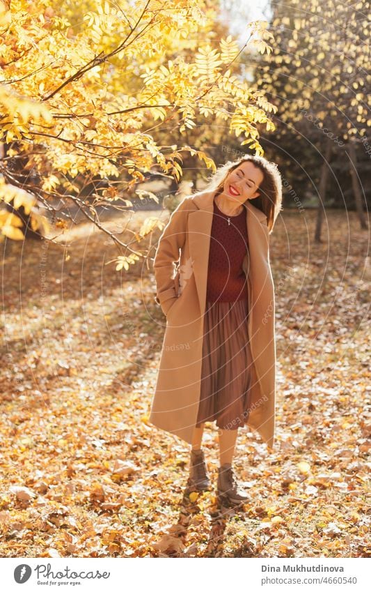Schöne Frau in beigem Mantel, weinrotem Pullover, braunem Rock und rotem Lippenstift Make-up, zu Fuß im Herbst Park. Porträt eines Mädchens im Herbst Natur mit gelbem Laub.