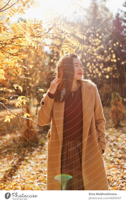 Schöne Frau in beigem Mantel, weinrotem Pullover, braunem Rock und rotem Lippenstift Make-up, stehend im Herbst Park. Porträt eines Mädchens in der herbstlichen Natur mit gelbem Laub.