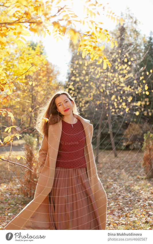 Schöne Frau in beigem Mantel, weinrotem Pullover, braunem Rock und rotem Lippenstift Make-up, stehend im Herbst Park. Porträt eines Mädchens in der herbstlichen Natur mit gelbem Laub.