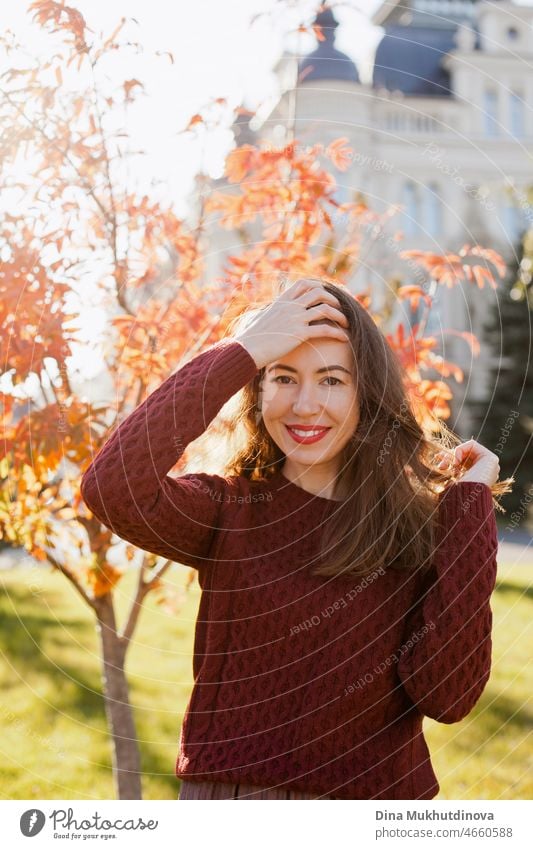 Junge Frau in weinrotem Pullover und rotem Lippenstift Make-up, stehend im Herbst Park in der Nähe historischer Gebäude. Porträt eines Mädchens im Herbst Park mit orange roten Laub.