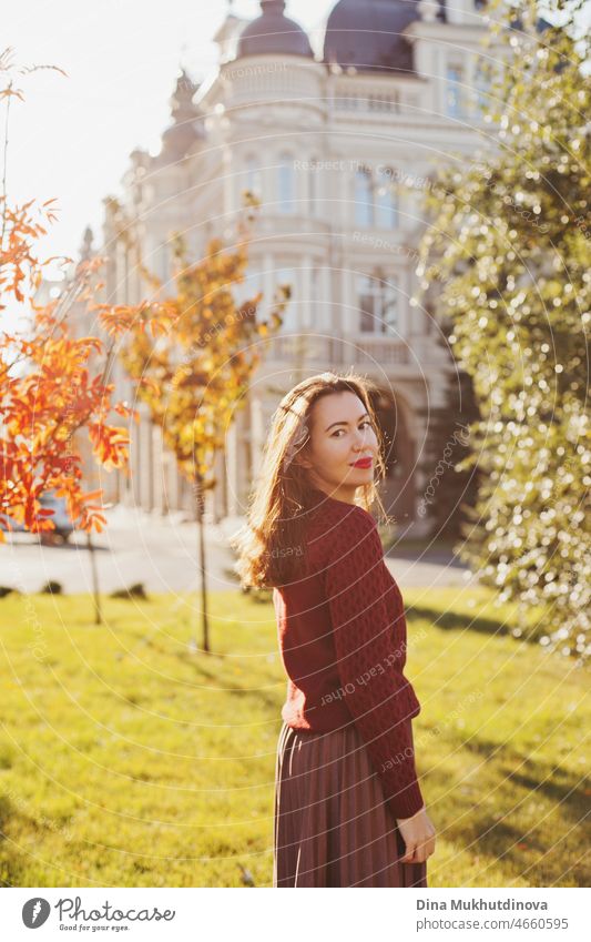 Junge Frau in weinrotem Pullover, braunem Rock und rotem Lippenstift Make-up, stehend im Herbst Park in der Nähe historischer Gebäude. Porträt eines Mädchens im Herbst Park mit orange-rotem Laub.
