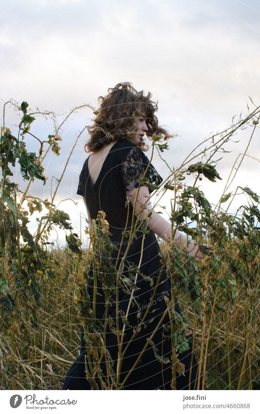 Junge Frau mit Locken steht in dunklem Kleid im Feld, vertrocknete Gräser um sie herum. Pflanze natürlich Außenaufnahme Ganzkörperaufnahme Tag Mädchen Natur