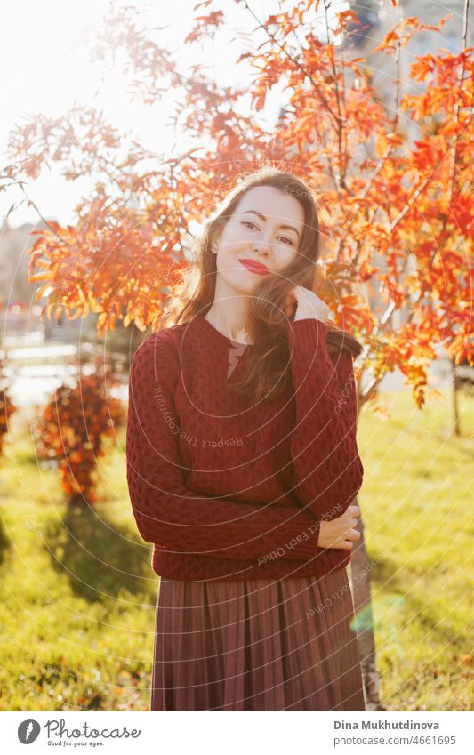 Junge Frau in weinrotem Pullover, braunem Rock und rotem Lippenstift Make-up, stehend in der Nähe von Herbstbaum. Porträt eines Mädchens im Herbst Park mit orange roten Laub.