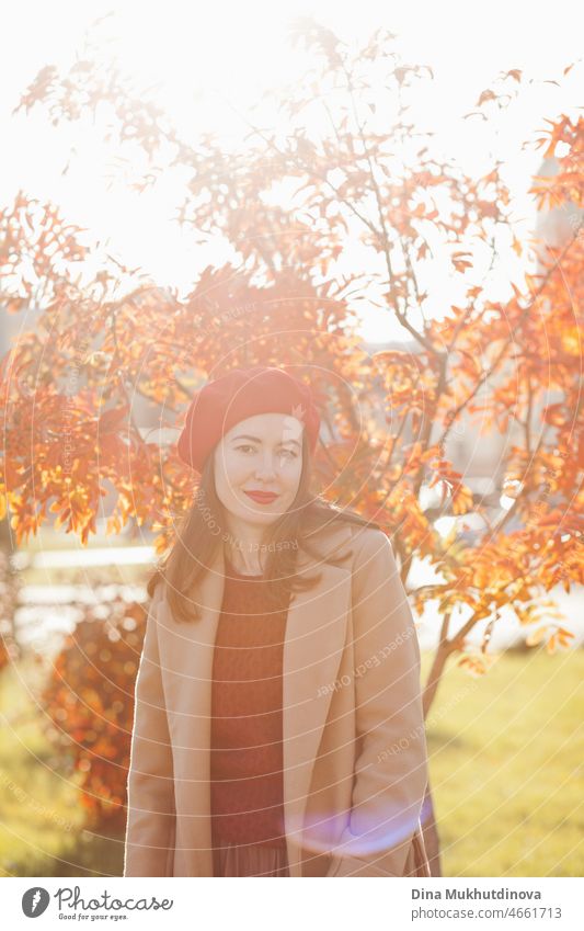 Junge Frau mit roter Baskenmütze, beigem Mantel und weinrotem Pullover und rotem Lippenstift, die in der Nähe eines Herbstbaums steht. Porträt eines Mädchens im Herbst Park mit orange roten Laub.