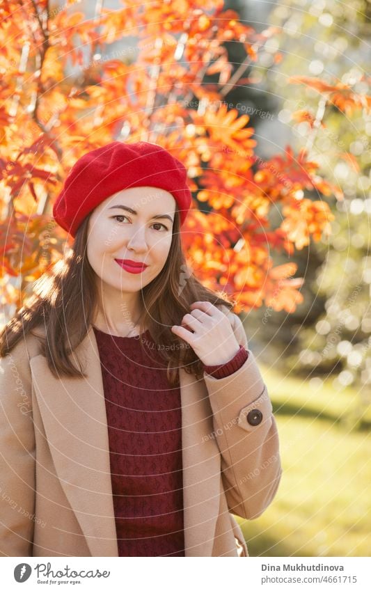 Junge Frau mit roter Baskenmütze, beigem Mantel und weinrotem Pullover und rotem Lippenstift, die in der Nähe eines Herbstbaums steht. Porträt eines Mädchens im Herbst Park mit orange roten Laub.