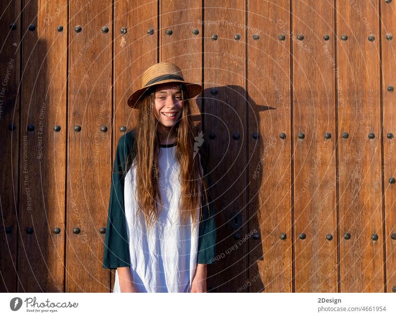 Junge glückliche Teenagerin mit Sommerkleidung und Hut, die gegen eine alte Holztür im Freien steht und in die Kamera schaut Glück Mädchen Stehen Lächeln