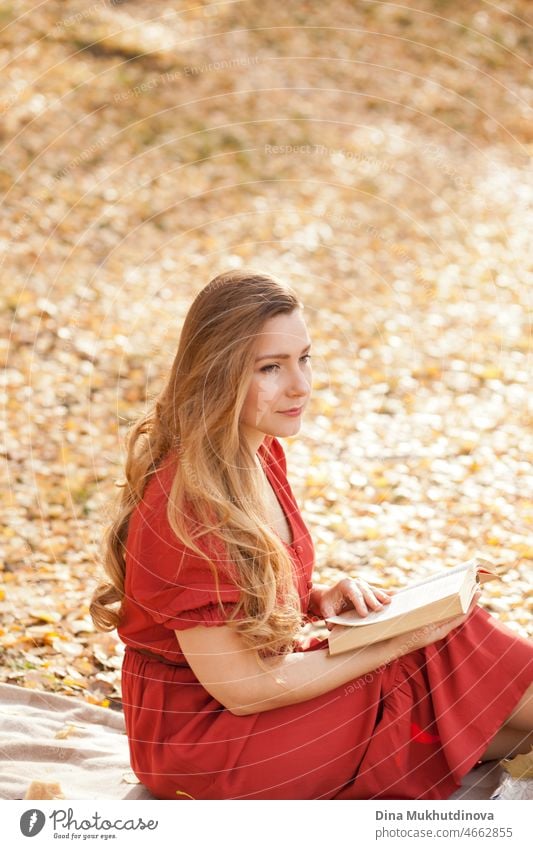 Schöne Frau mit langen Haaren in rotem Kleid Lesen eines Buches im Herbst Park im Freien in der Natur. Romantische Buchliebhaberin liest draußen an einem sonnigen Herbsttag.