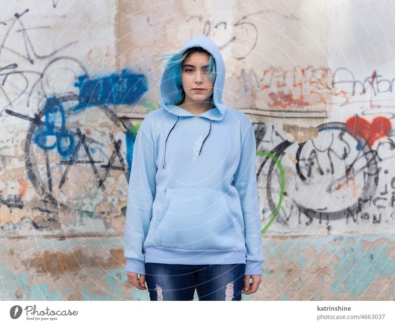 Blauhaariges Teenager-Mädchen in hellblauem Oversize-Kapuzenpulli, das gegen eine Graffiti-Wand zeigt hell-blau Attrappe Übergröße Jeanshose blauhaarig