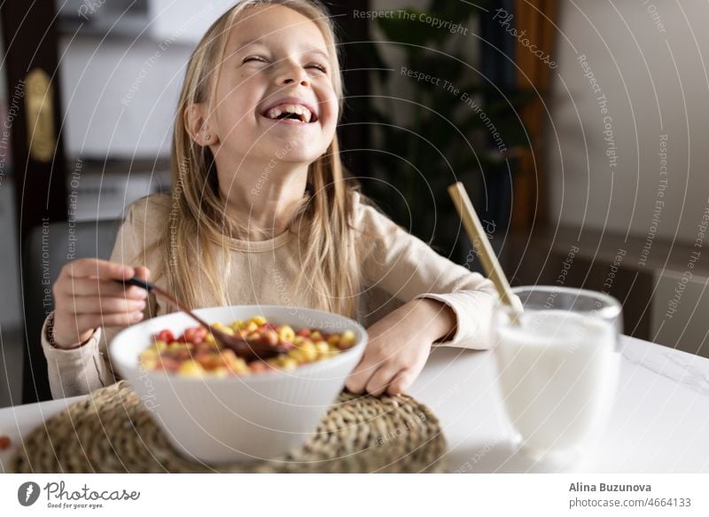 Nettes kleines kaukasisches Mädchen sitzt am Tisch in der Küche am frühen Morgen und bereitet Frühstück mit bunten Cornflakes und Milch. Kind genießt das Leben mit gesunden Lebensmitteln, gesunden Lebensstil Konzept