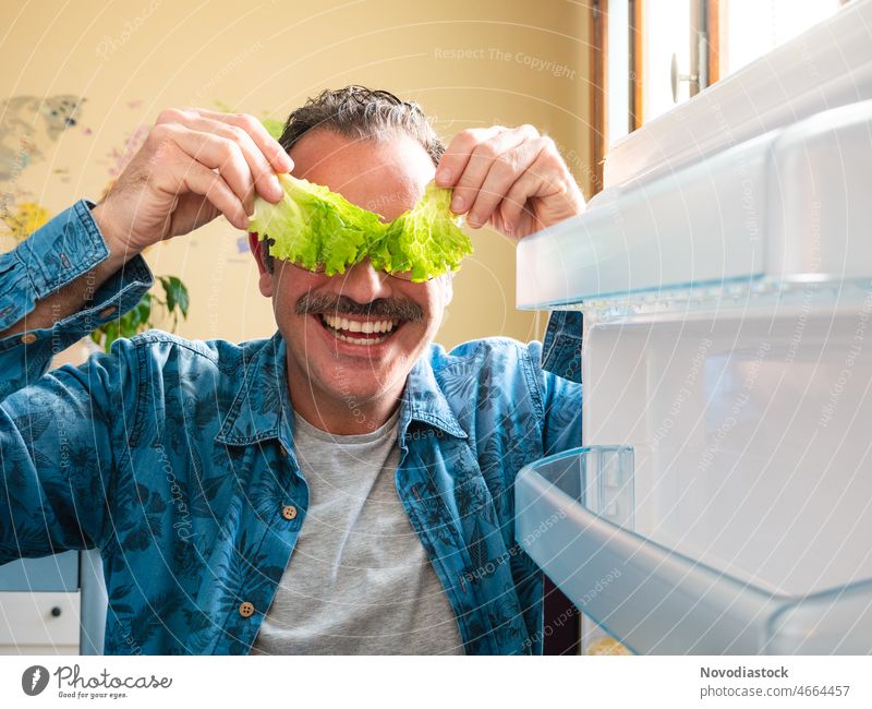 Porträt eines Mannes mittleren Alters, der seine Augen mit zwei Salatblättern bedeckt, die Kühlschranktür geöffnet hat und ein komisches Gesicht macht positiv